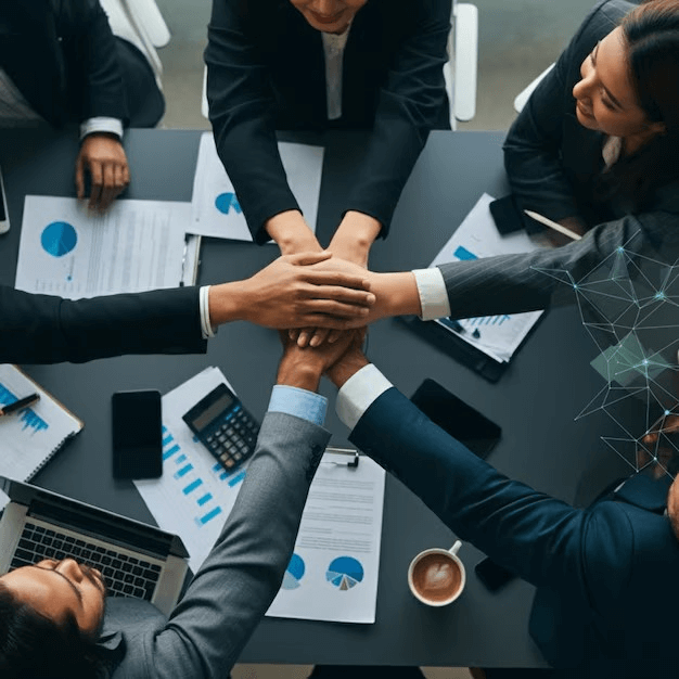 A top-down view of hands stacked together on a conference table surrounded by documents, laptops, and a coffee cup, symbolizing teamwork.